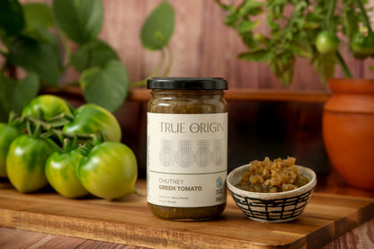 A jar of True Origin Green Tomato Chutney sitting on a wooden surface with a bowl of green tomato chutney and some green tomatoes.