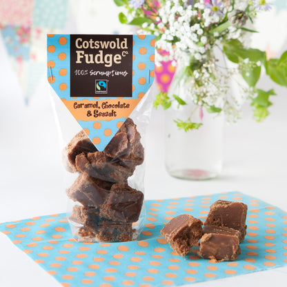 A bag of 100% scrumptious Fairtrade Caramel, Chocolate and Seasalt Fudge made by Cotswold Fudge Co. The bag is transparent with chunks of fudge on display.  The bag is sitting on a light blue napkin with orange polkadots with some flowers in the background.