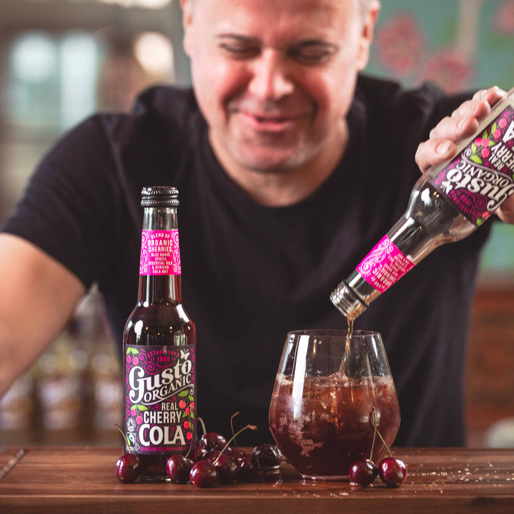Gusto Organic Cherry Cola being poured into a glass with ice next to an unopened bottle.