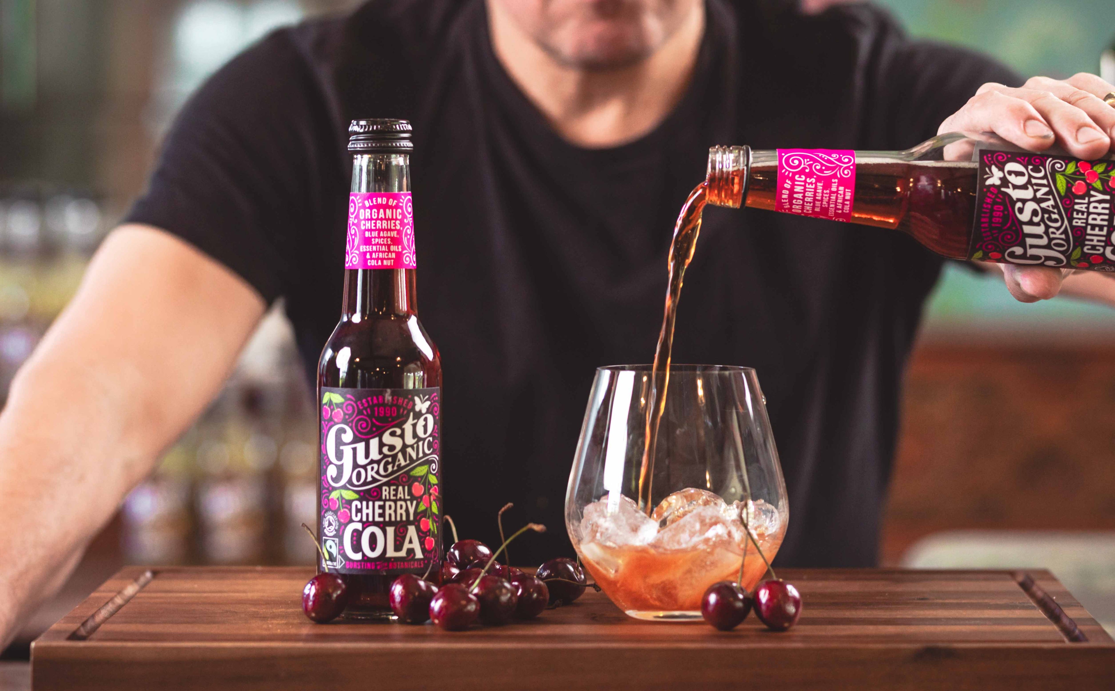 Gusto Organic Fairtrade Real Cherry Cola being poured into a glass with ice next to an unopened bottle.