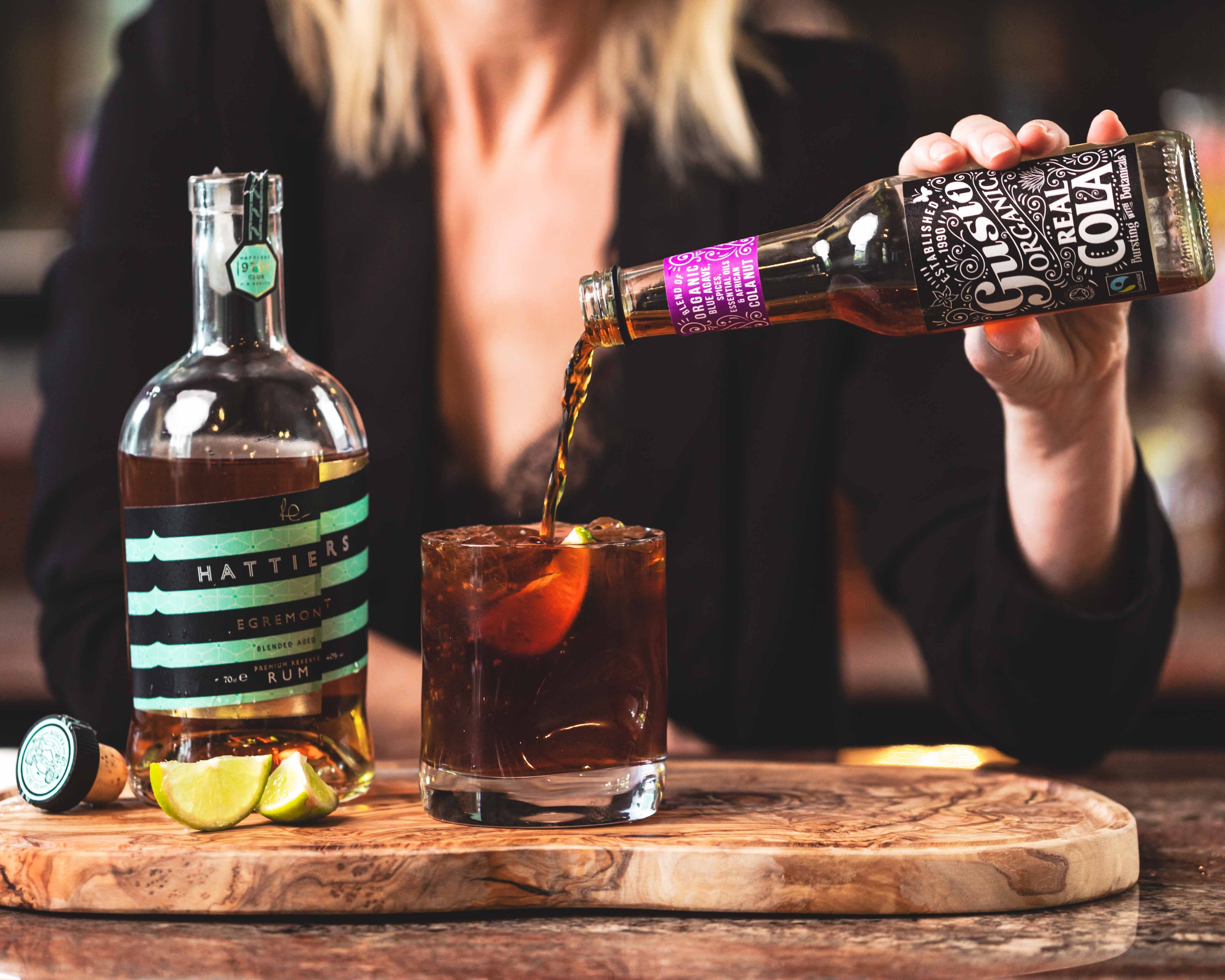 A woman sitting at a table pouring some Gusto organic real cola into a glass with rum and ice.