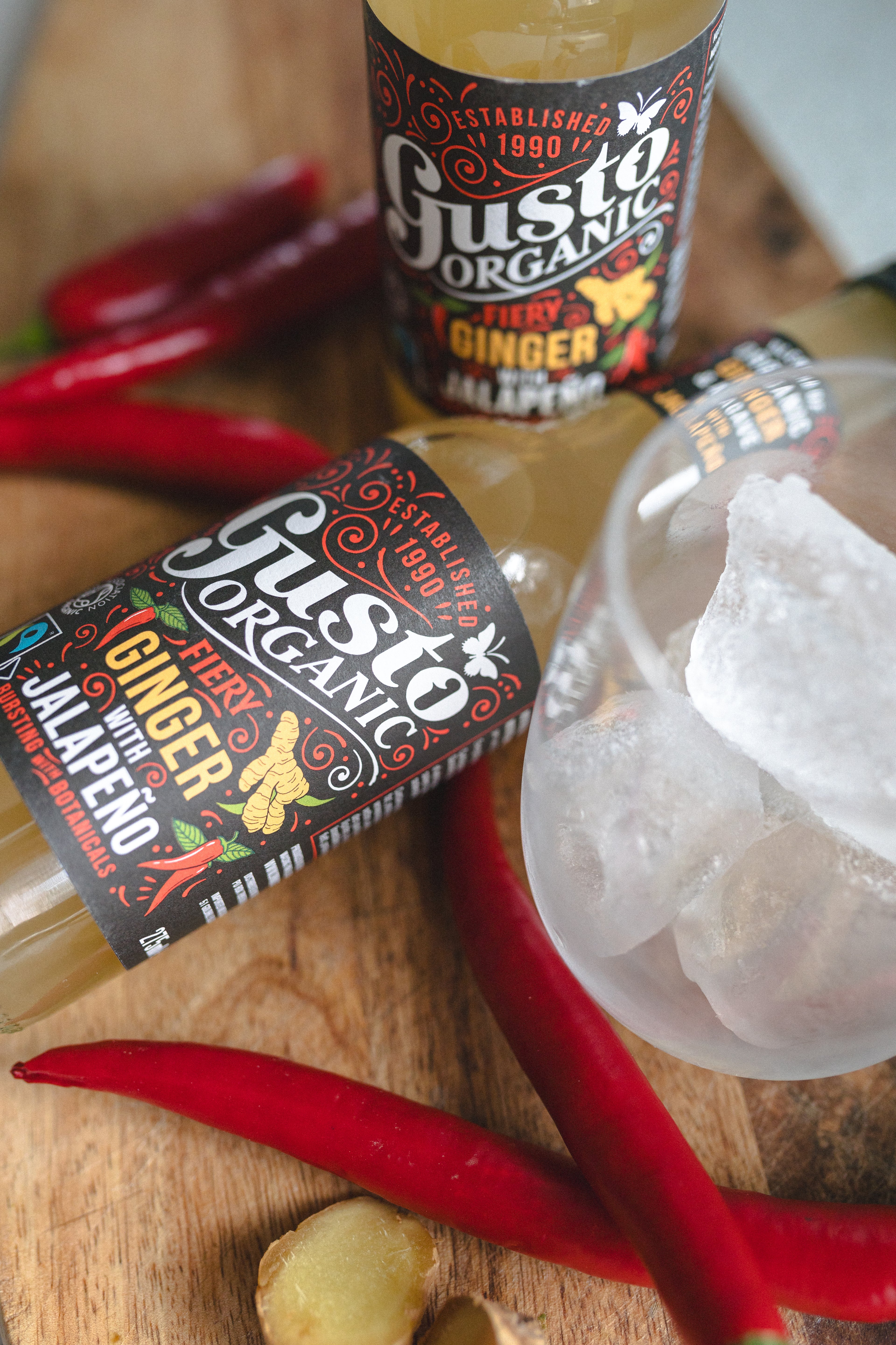 Two bottles of Fairtrade Gusto Organic Ginger with Jalapeno soft drink on a wooden surface surrounded with chillies and a glass with ice.