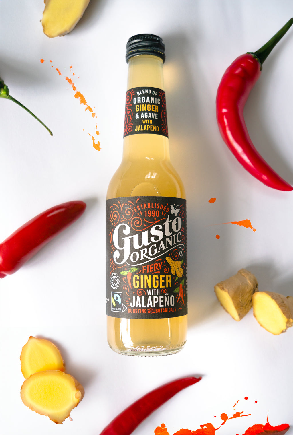 A bottle of Fairtrade Gusto Organic Ginger with Jalapeno soft drink on a white surface surrounded with chillies and a ginger.