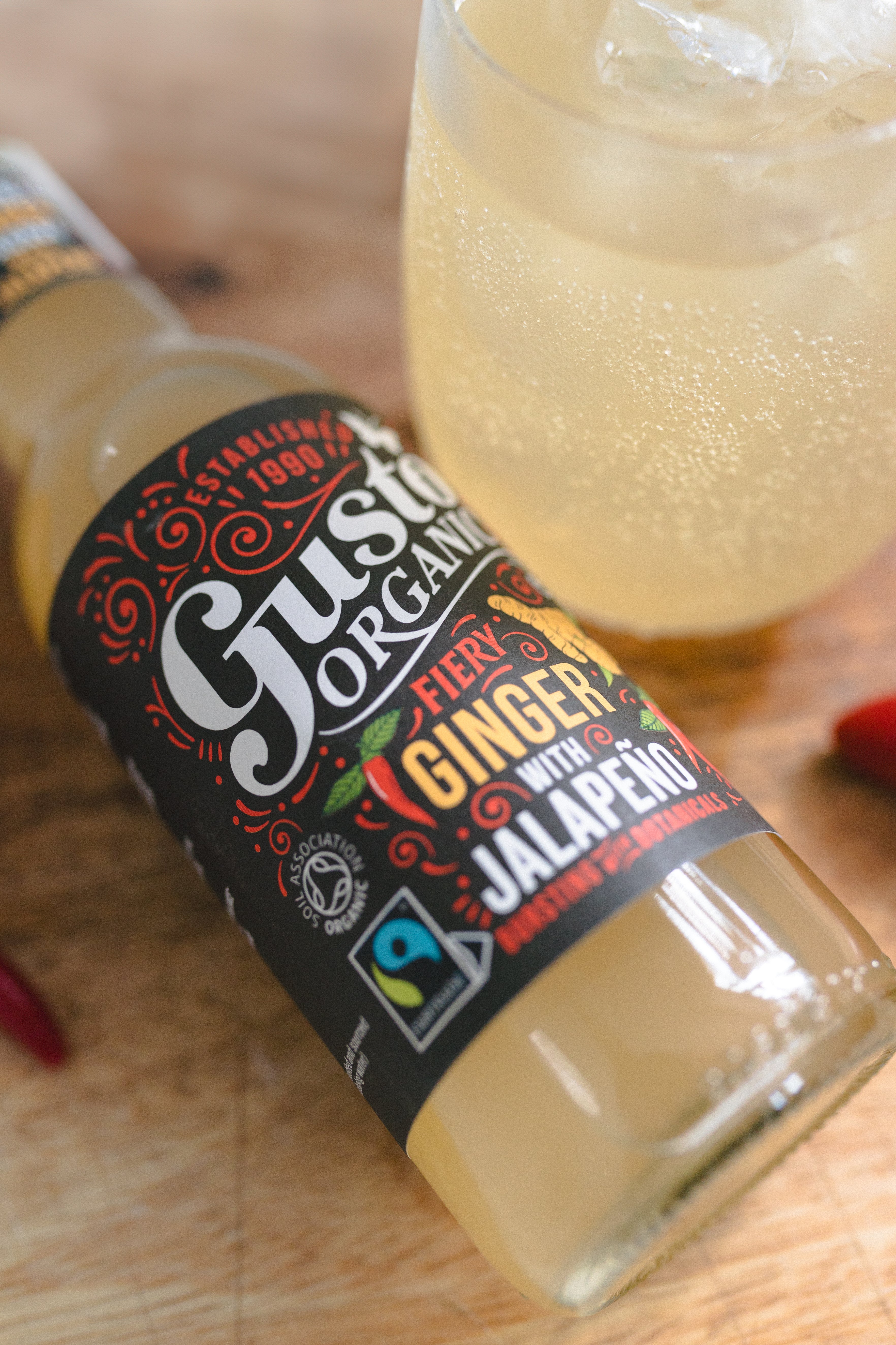 A bottle of Fairtrade Gusto Organic Ginger with Jalapeno soft drink on a wooden surface surrounded with chillies and a glass with ice.