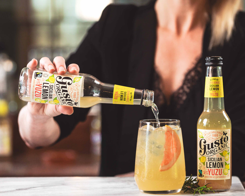 A woman pouring some Gusto Organic Sicilian Lemon with Yuzu into a glass with ice next to an unopened bottle.