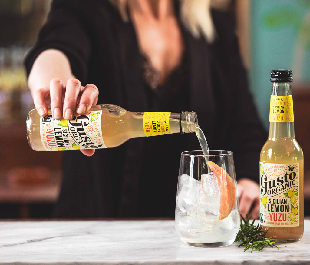 A woman pouring Gusto Organic Sicilian Lemon with Yuzu into a glass with ice with an unopened bottle beside the glass.