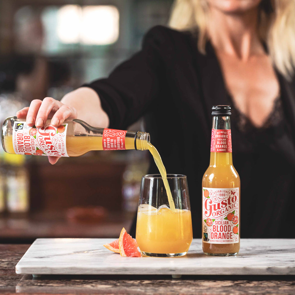 Bottle of Fairtrade Gusto Organic Sicilian Blood Orange being poured into a glass with ice next to an unopened bottle.
