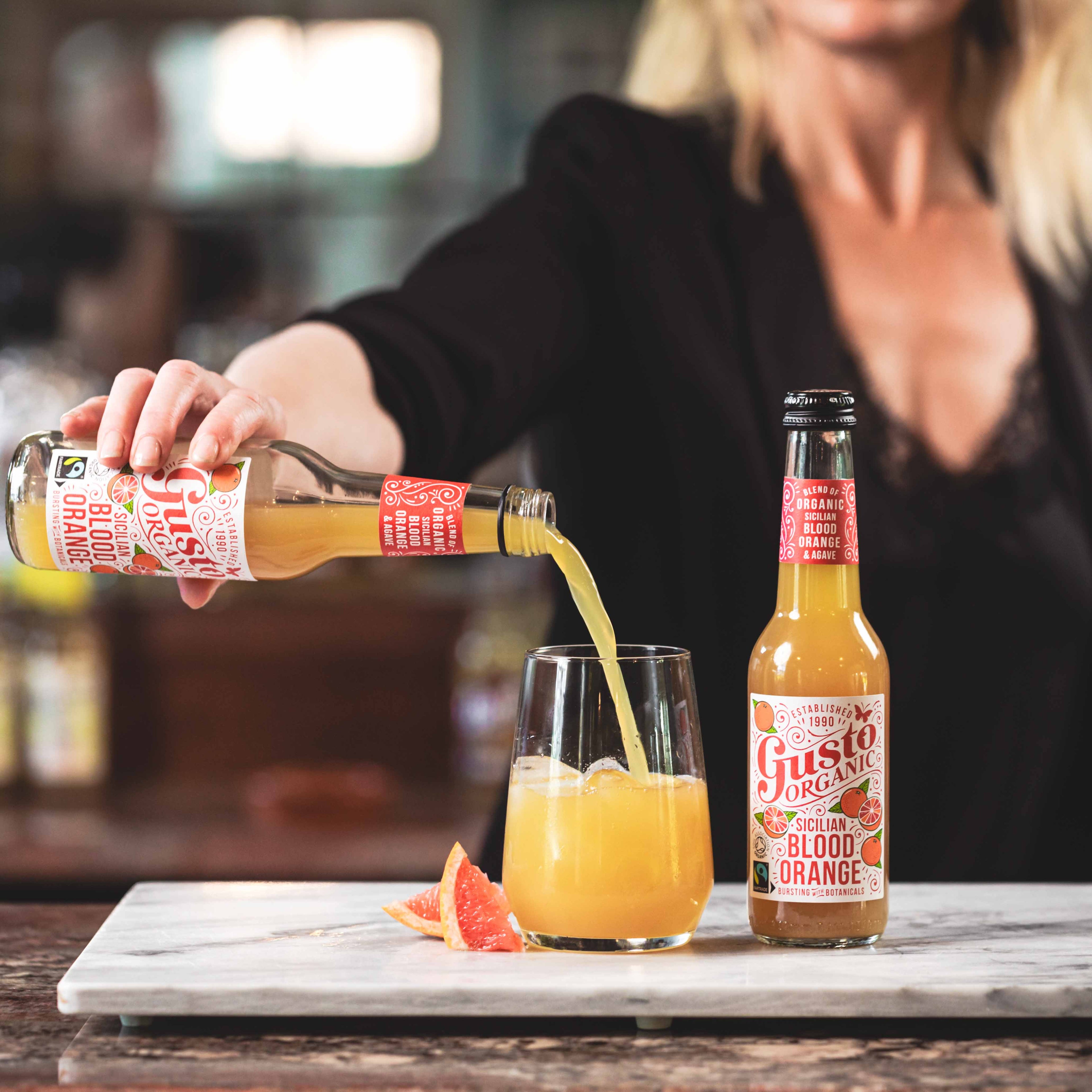 Bottle of Fairtrade Gusto Organic Sicilian Blood Orange being poured into a glass with ice next to an unopened bottle.