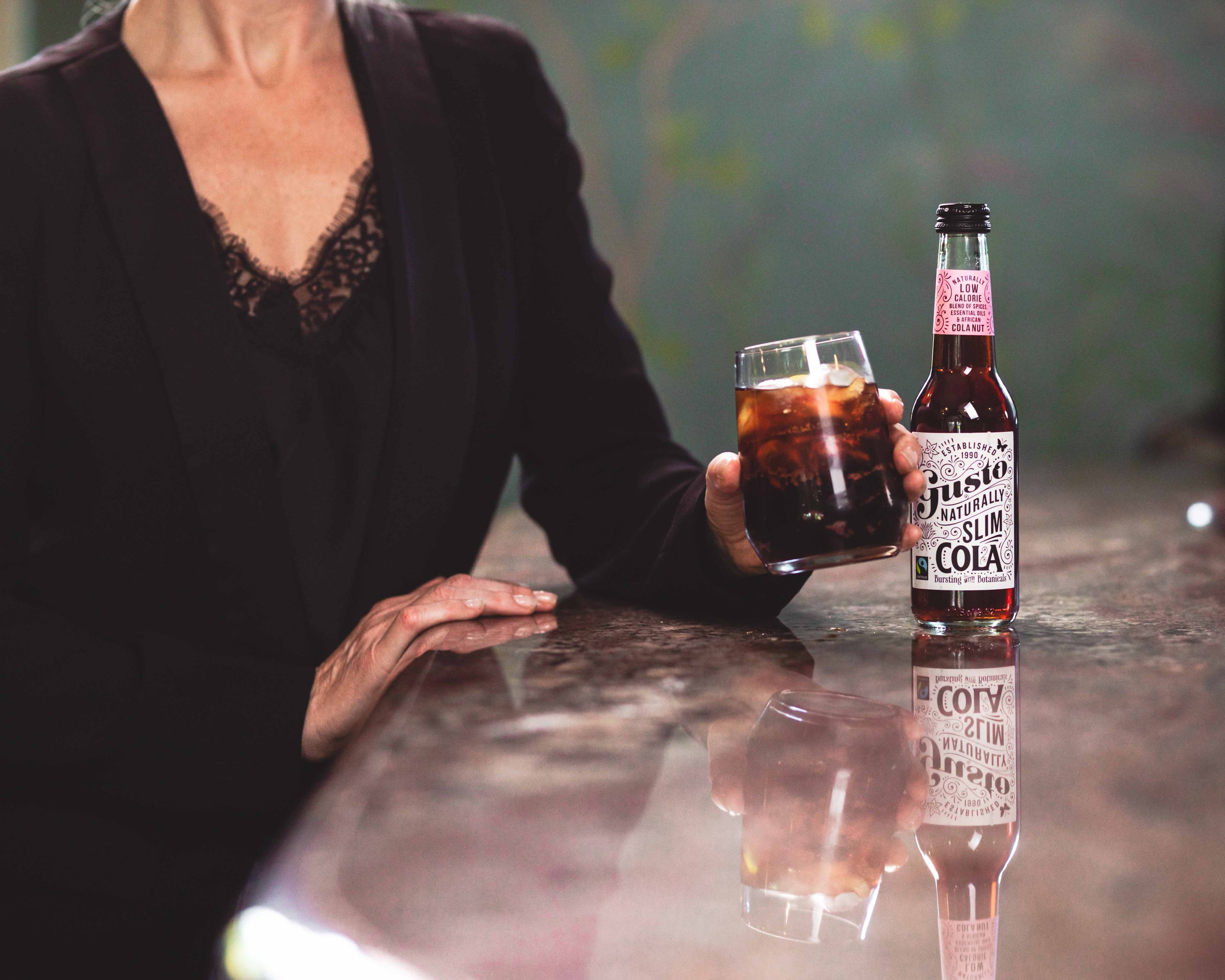 A woman holding Gusto Organic Slim Cola in a glass with ice.
