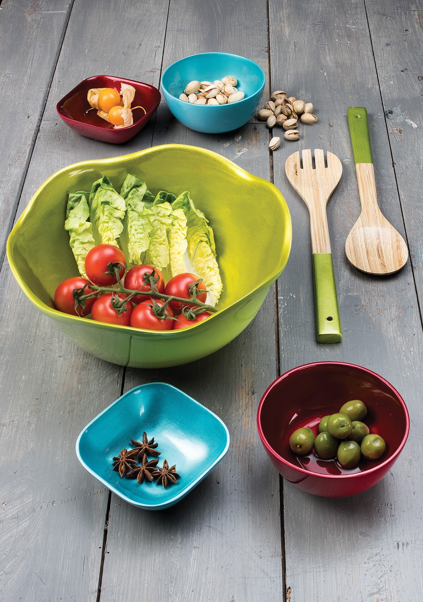 A lime green salad bowl containing lettuce and vine tomatoes in the middle next to a range of smaller blue and red round and square bowls containing fruit, nut and spices.