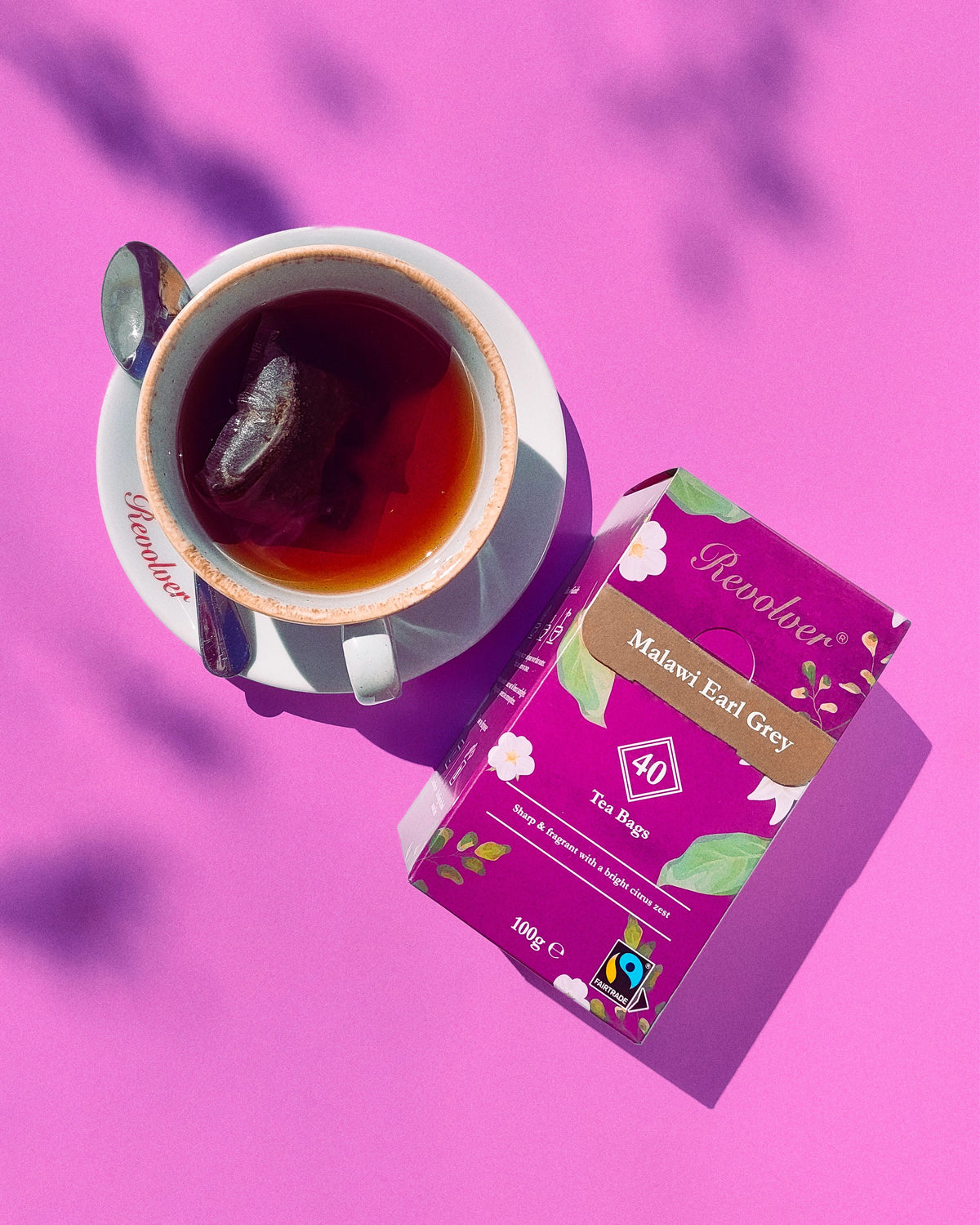 A cup of Earl Grey tea on a saucer beside a purple tea box labeled Revolver Malawi Earl Grey, arranged on a bright pink background with leaf shadows.