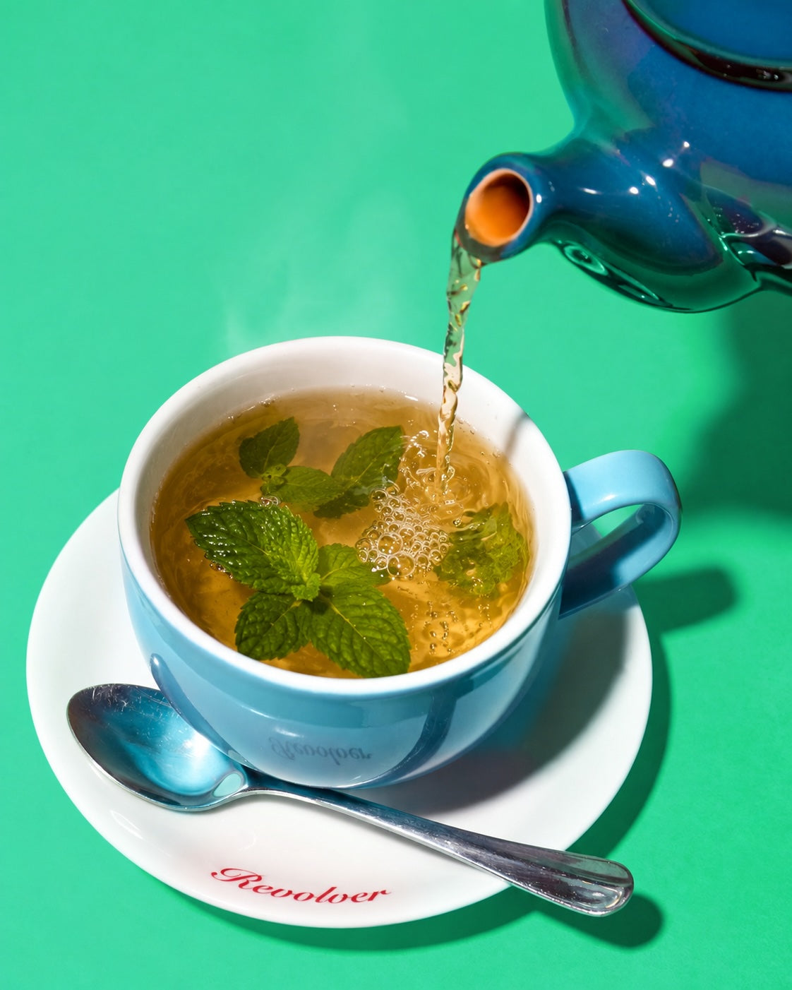 A blue teapot pours amber tea into a matching blue teacup on a white saucer with a silver spoon, set against a bright green background. The cup has mint leaves inside, and the saucer shows the brand name 'Revolver'.