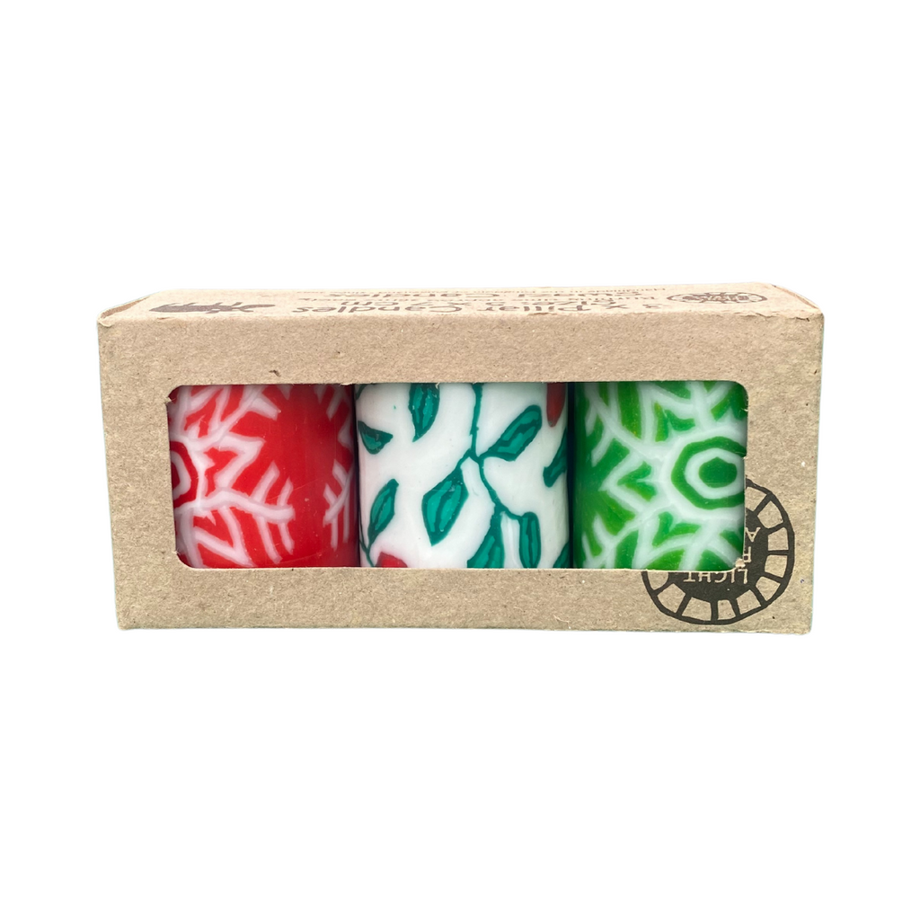 A triple pack of church candles with a Christmas design.  The is a red candles with a white snowflake design, a white candle with a holly design and a green candle with a white snowflake design.