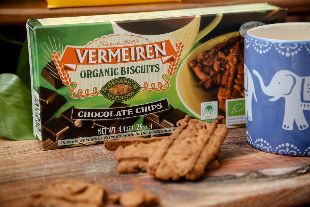 A packet of Vermeiren organic chocolate chip biscuits next to a blue mug with an elephant design and some loose speculoos biscuits on a chopping board.