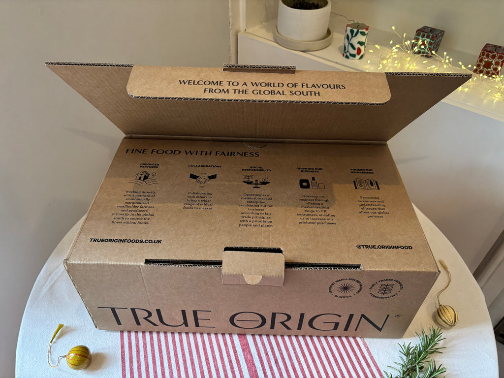 Open cardboard hamper box with 'True Origin' branding on a table with decorative items.