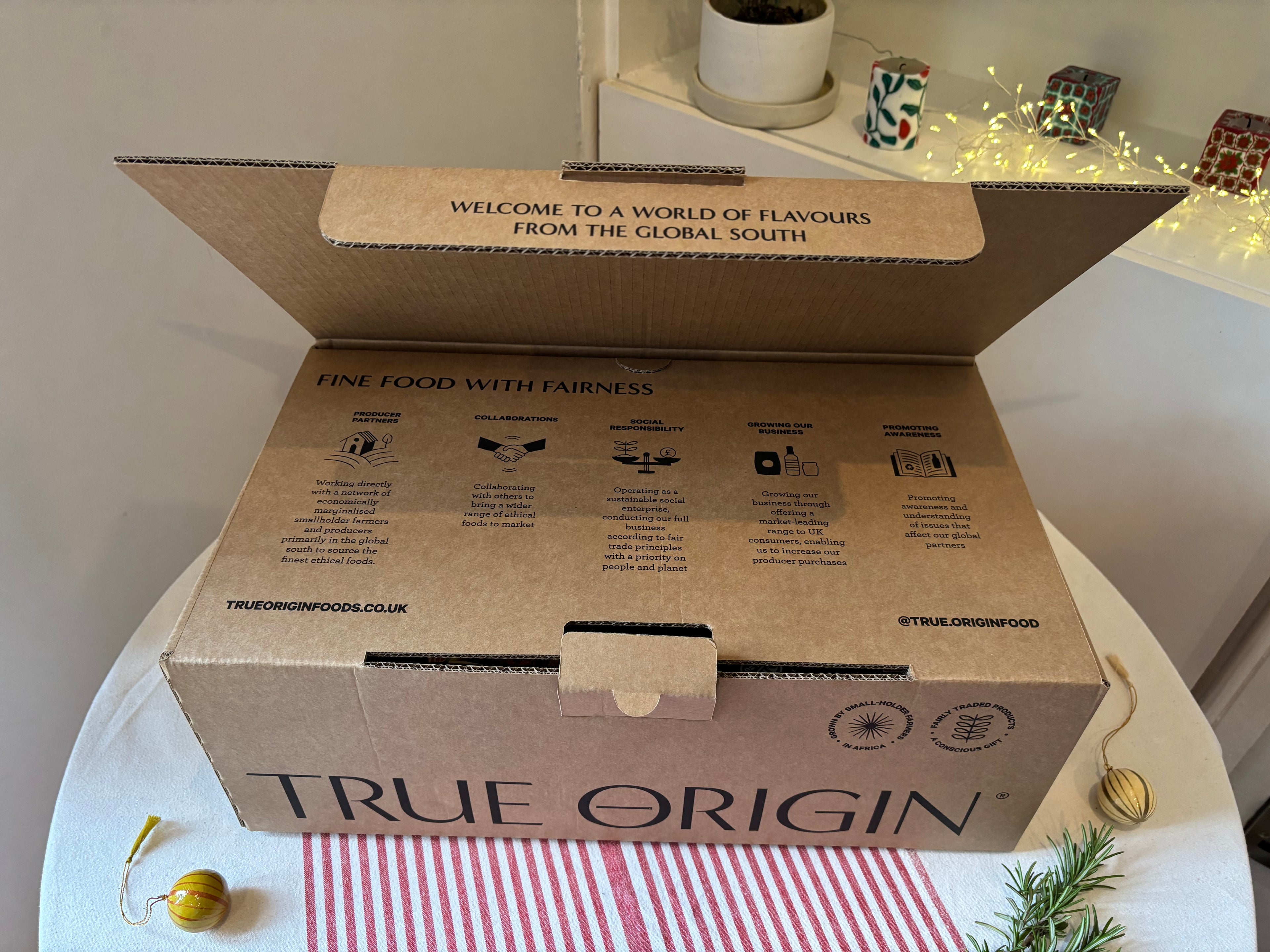 Open cardboard hamper box with 'True Origin' branding on a table with decorative items.