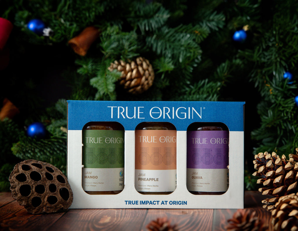 A gift set box containing three jars of True Origin brand jams: Guava Jam, Pineapple Jam, and Mango Jam, each with a clear label showing the flavour and 'True Impact at Origin' text.  The gift set is surrounded by festive greenery and pine cones.