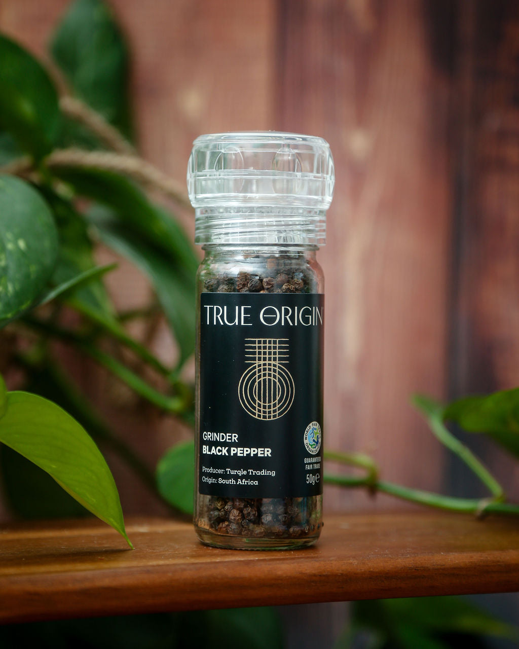 A small glass bottle of True Origin Black Pepper with a built in grinder on a wooden chopping board with some green leaves in the background.