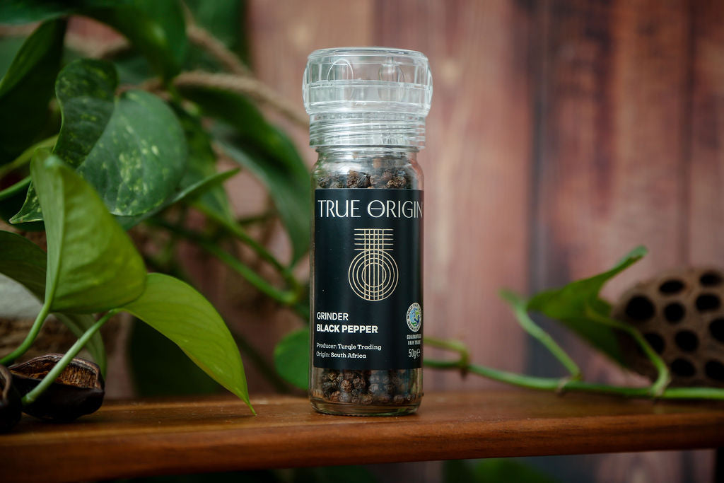 A small glass bottle of True Origin Black Pepper on a wooden chopping board with green leaves in the background.