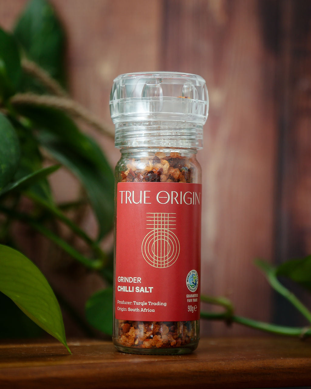 A red glass jar of chilli salt with a built in grinder on a wooden shelf with some green leaves in the background.
