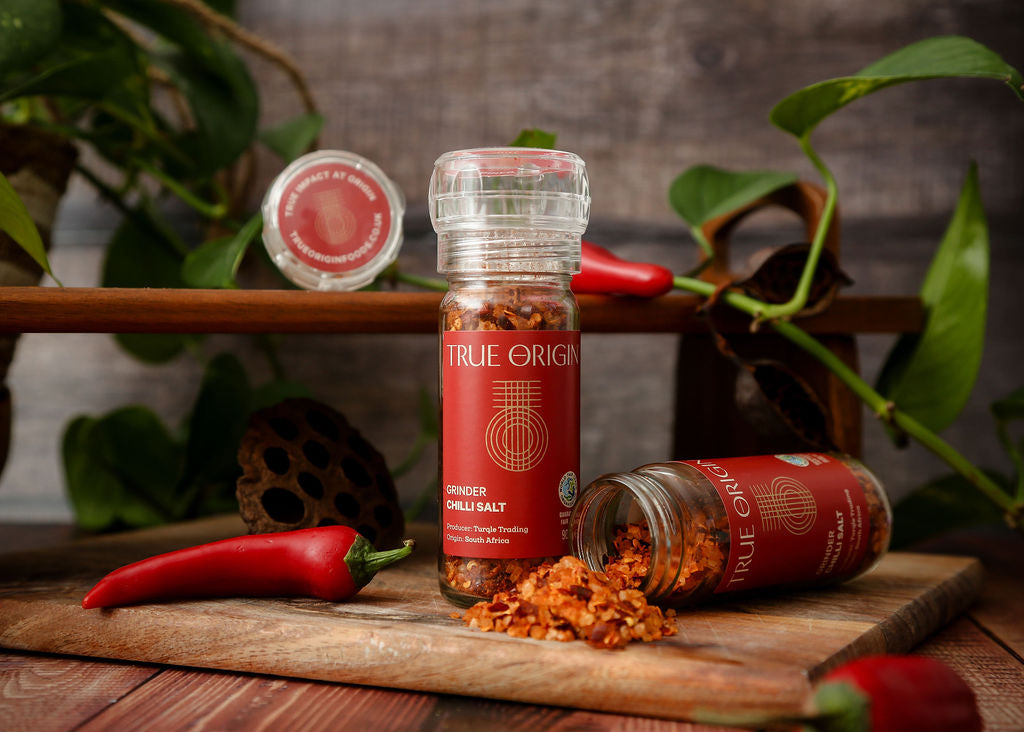 A red True Origin Chilli Salt grinder next to an open red glass jar of chilli salt lying on its side with fair trade chilli salt poured onto a chopping board next to some chillies and green leaves in the background.