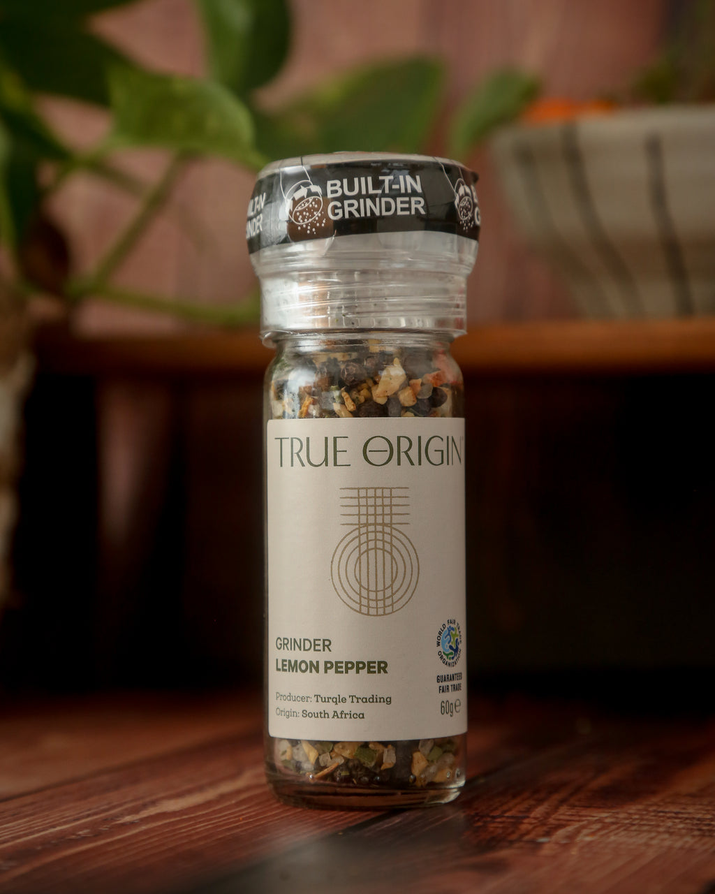 A clear plastic grinder bottle labelled 'True Origin' filled with a mixture of black pepper, lemon and orange peels, and herbs, indicating it is a lemon pepper seasoning product.
