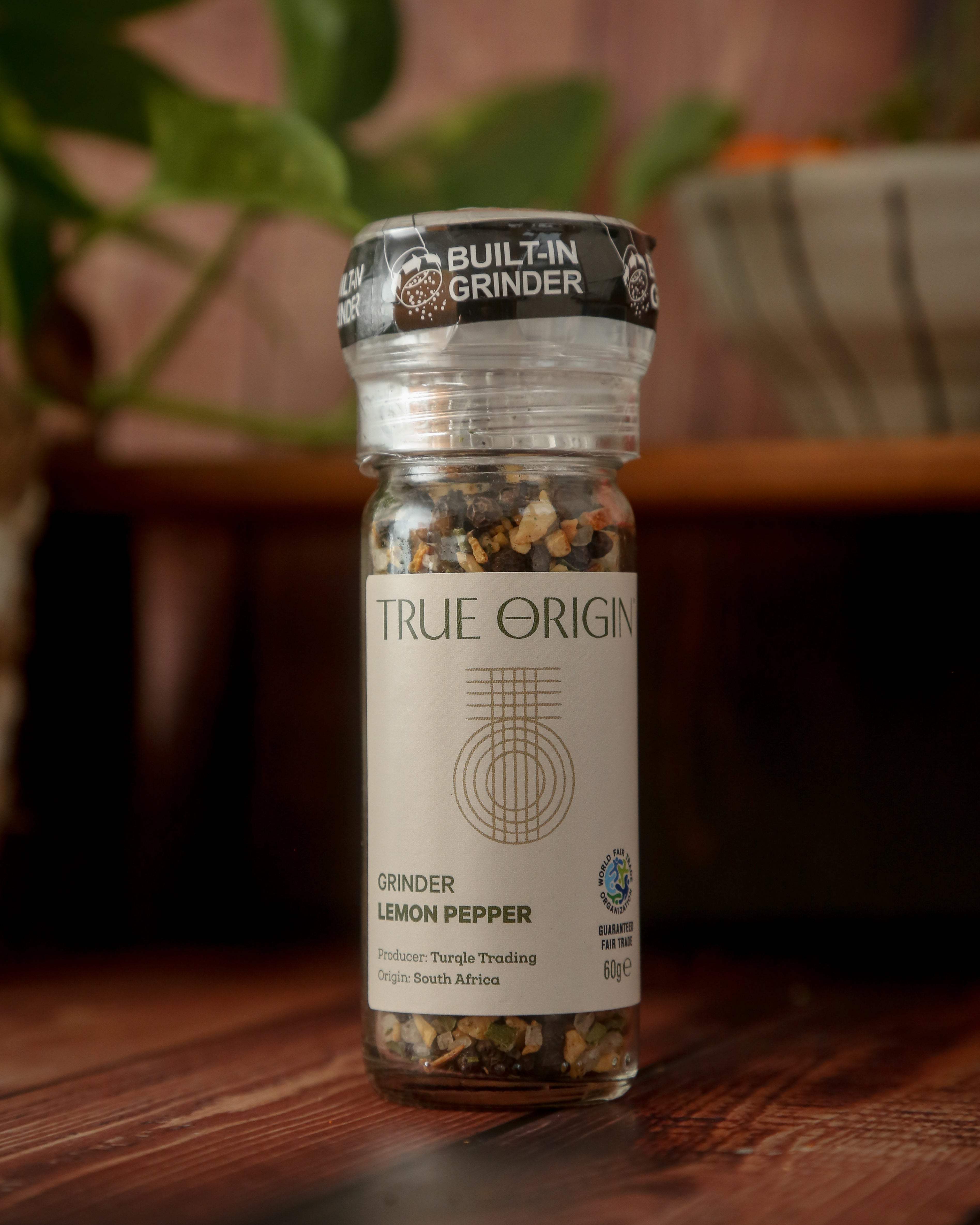 A clear plastic grinder bottle labelled 'True Origin' filled with a mixture of black pepper, lemon and orange peels, and herbs, indicating it is a lemon pepper seasoning product.