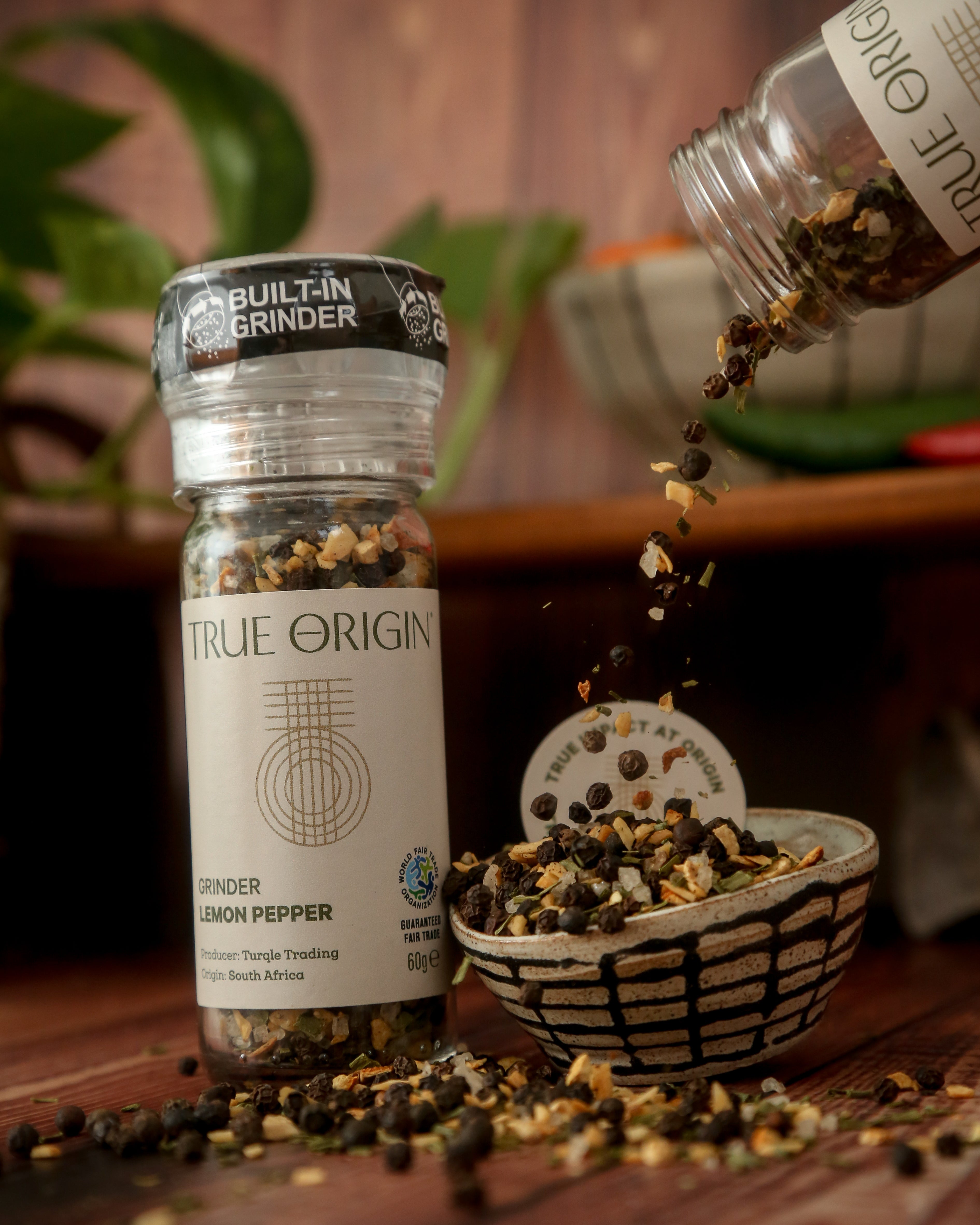A clear plastic grinder bottle labelled 'True Origin' filled with a mixture of black pepper, lemon and orange peels, and herbs, indicating it is a lemon pepper seasoning product.