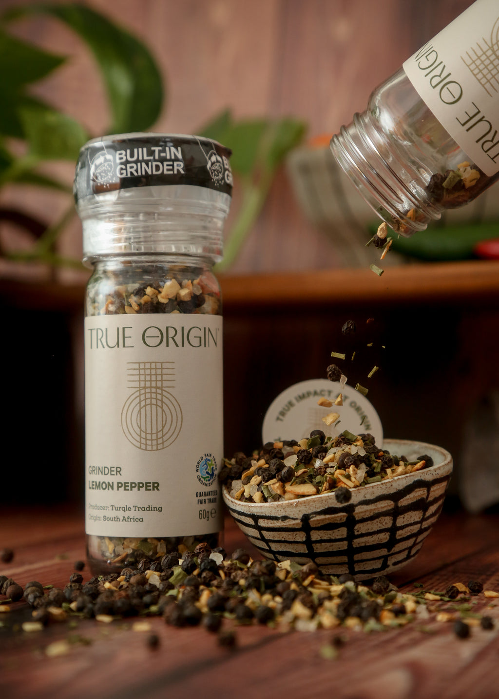 A clear plastic grinder bottle labelled 'True Origin' filled with a mixture of black pepper, lemon and orange peels, and herbs, indicating it is a lemon pepper seasoning product.