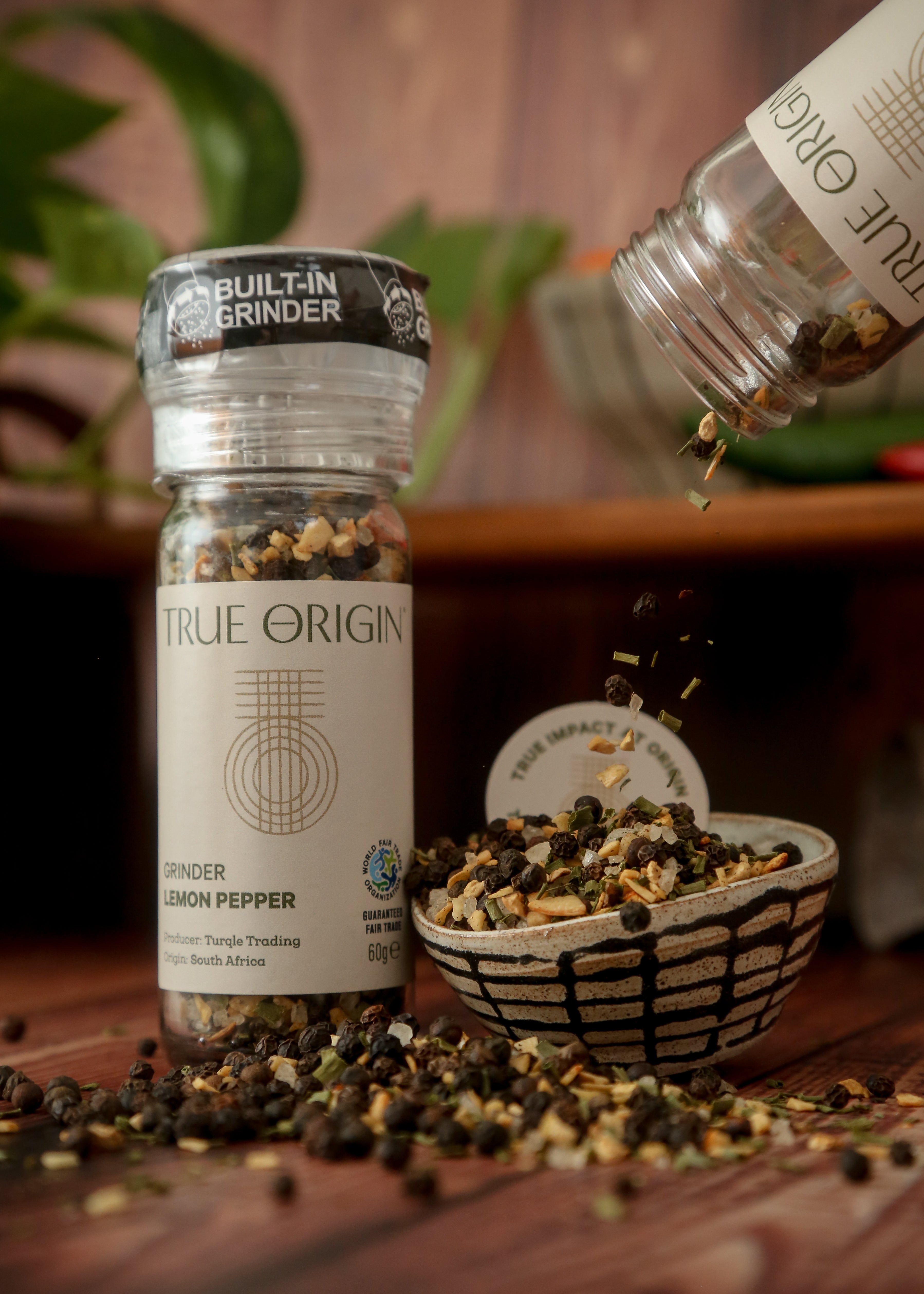 A clear plastic grinder bottle labelled 'True Origin' filled with a mixture of black pepper, lemon and orange peels, and herbs, indicating it is a lemon pepper seasoning product.