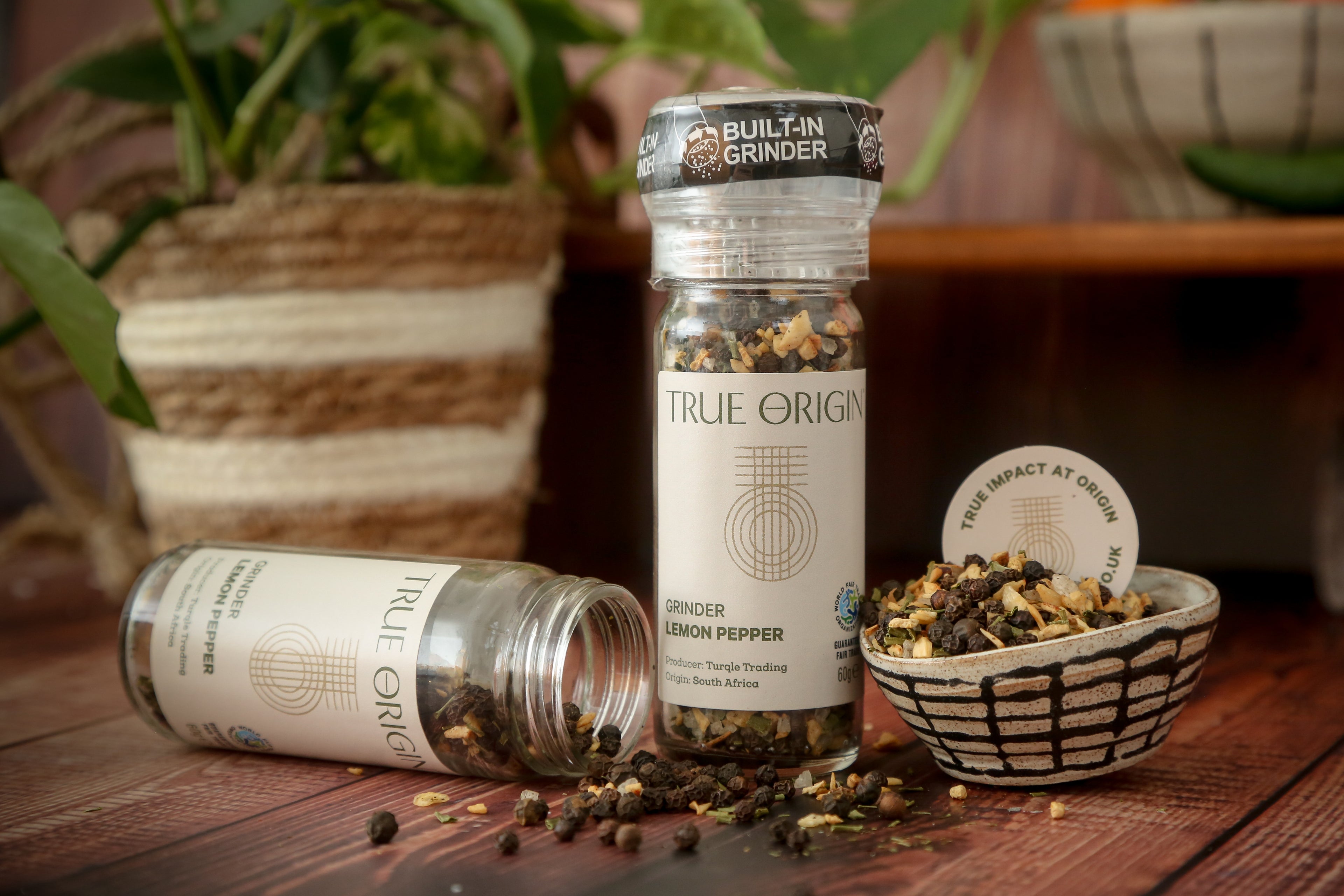 A clear plastic grinder bottle labelled 'True Origin' filled with a mixture of black pepper, lemon and orange peels, and herbs, indicating it is a lemon pepper seasoning product.