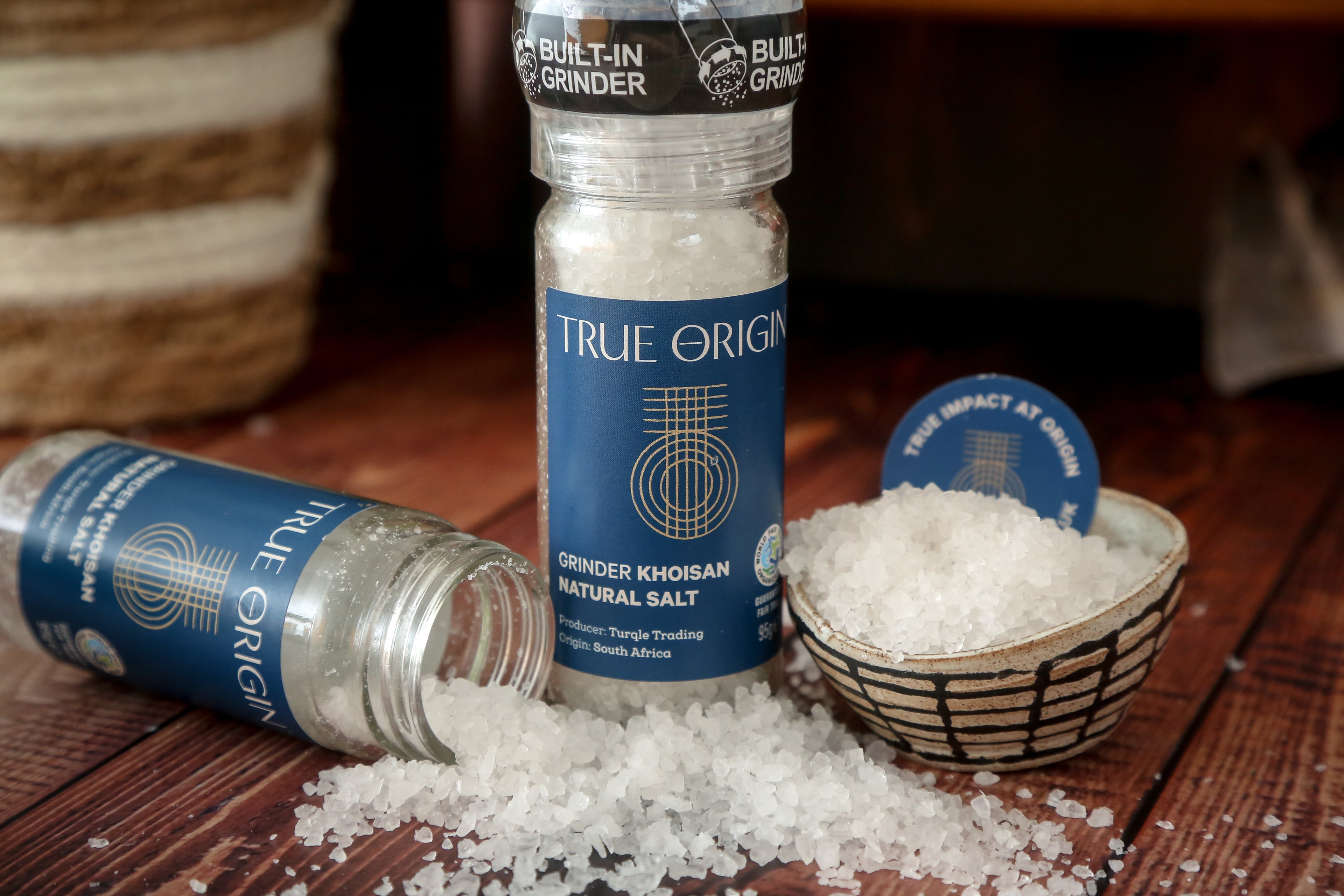 A clear glass bottle with a built-in grinder containing natural Khoisan salt, labelled 'True Origin'.  There is an opened jar of salt crystals being poured in a bowl and on the worktop.