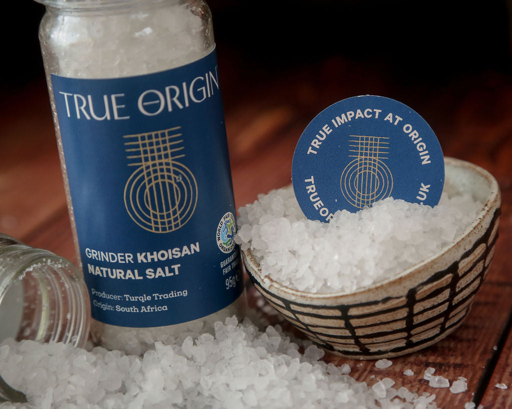 An open jar of True Origin khoisan natural salt from South Africa with a blue label next to a bowl overflowing with salt crystals and a True Impact at origin round logo.