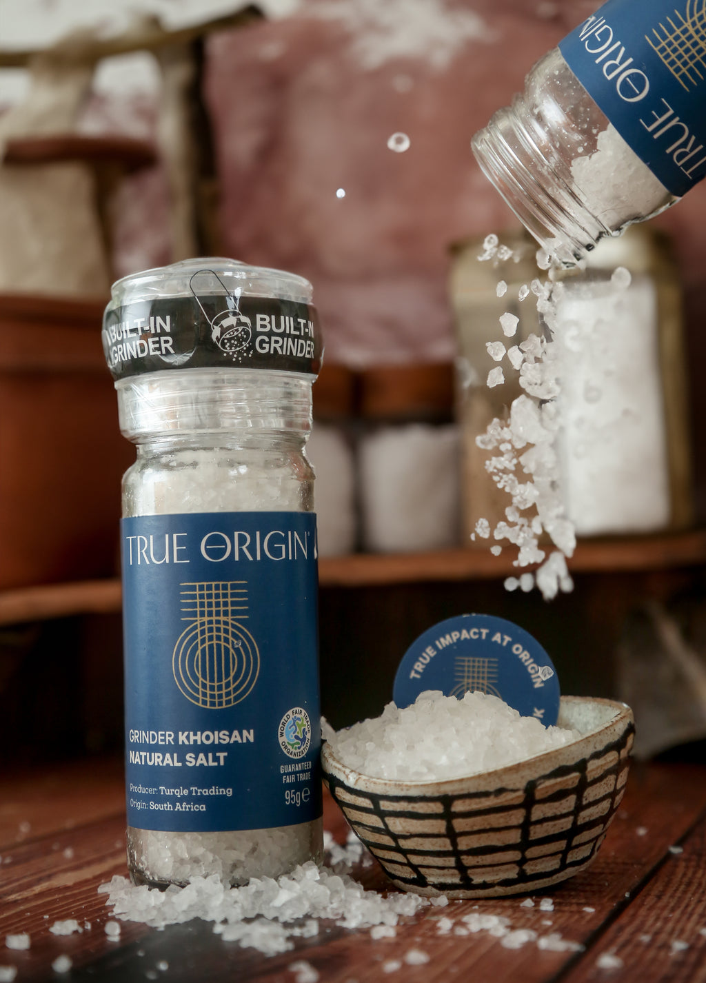 A clear glass bottle with a built-in grinder containing natural Khoisan salt, labelled 'True Origin'.  There is an opened jar of salt crystals being poured into a bowl.