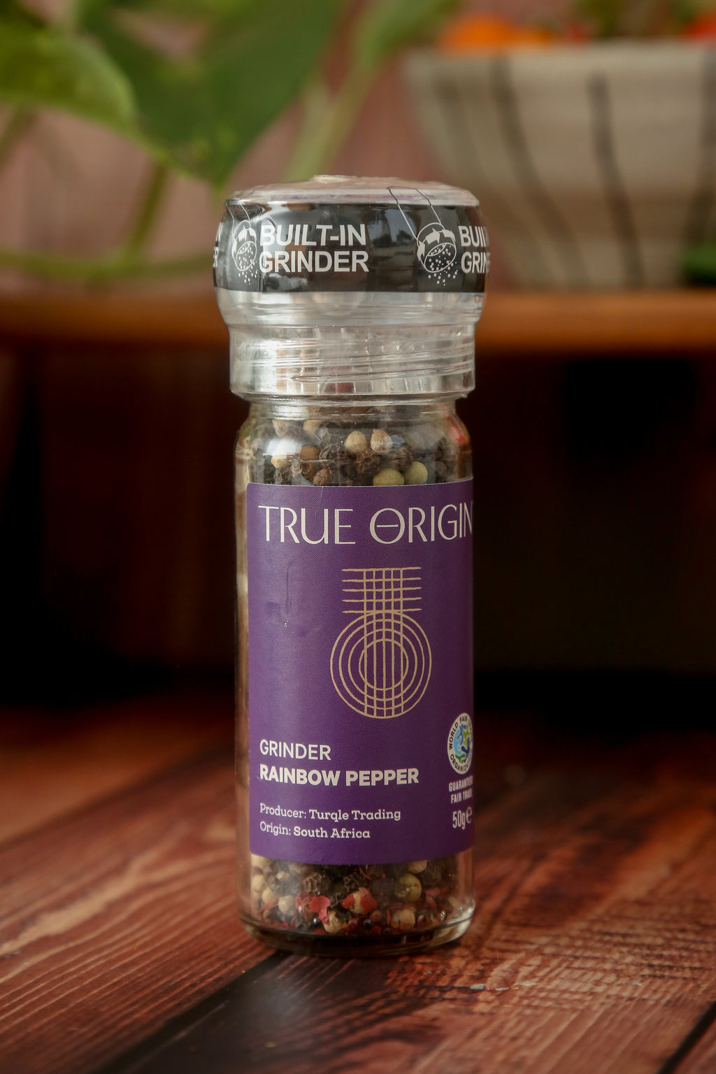 A clear glass pepper grinder with a built-in mechanism, containing a mix of black, white, green, and red pepper grains. The label shows 'True Origin' and indicates the product is a Rainbow Pepper Grinder, 50g size.