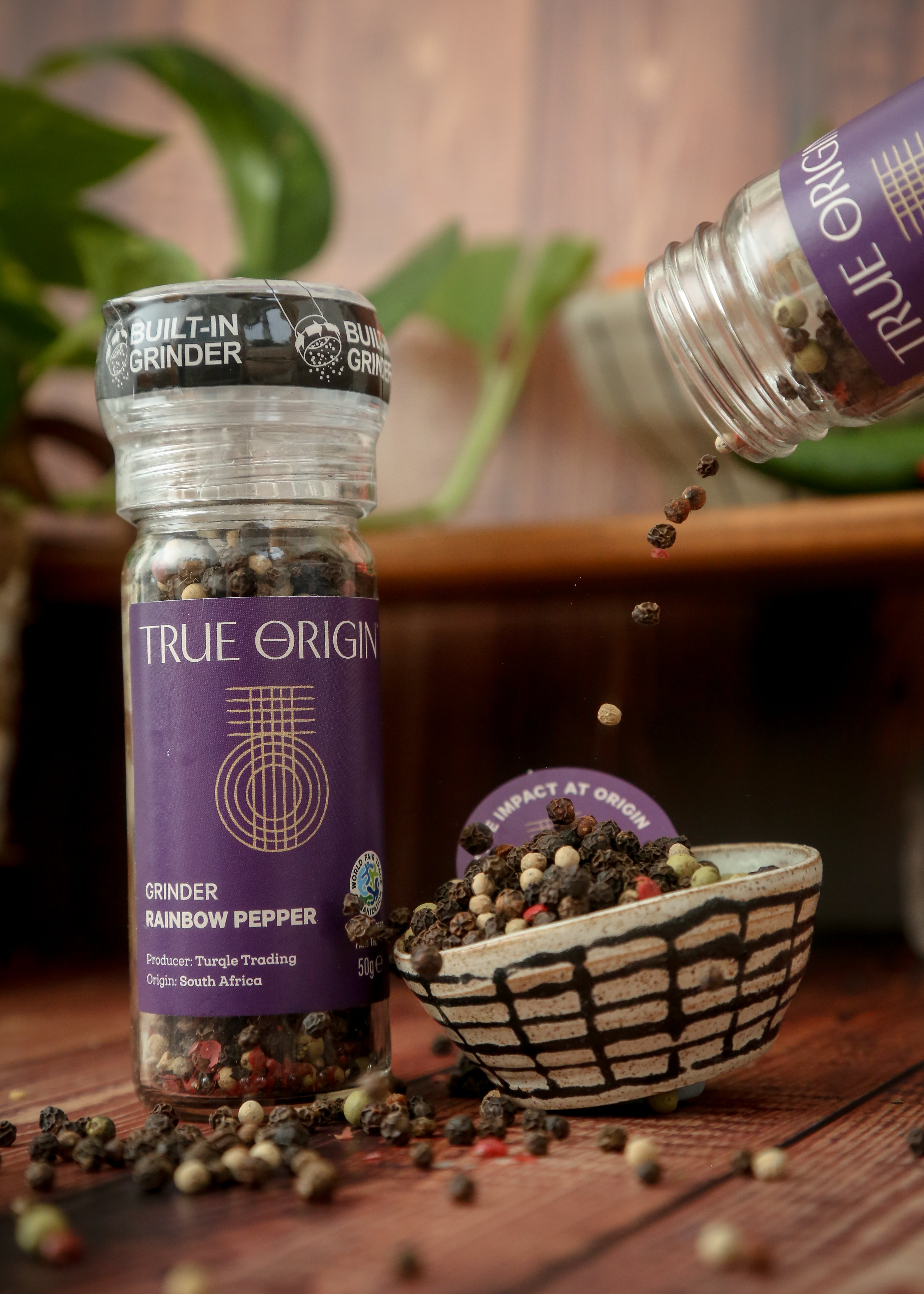 A clear glass pepper grinder with a built-in mechanism, containing a mix of black, white, green, and red pepper grains. The label shows 'True Origin' and indicates the product is a Rainbow Pepper Grinder, 50g size.