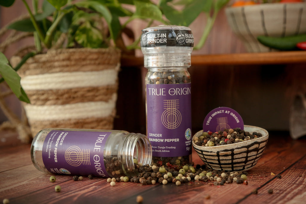 A clear glass pepper grinder with a built-in mechanism, containing a mix of black, white, green, and red pepper grains. The label shows 'True Origin' and indicates the product is a Rainbow Pepper Grinder, 50g size.