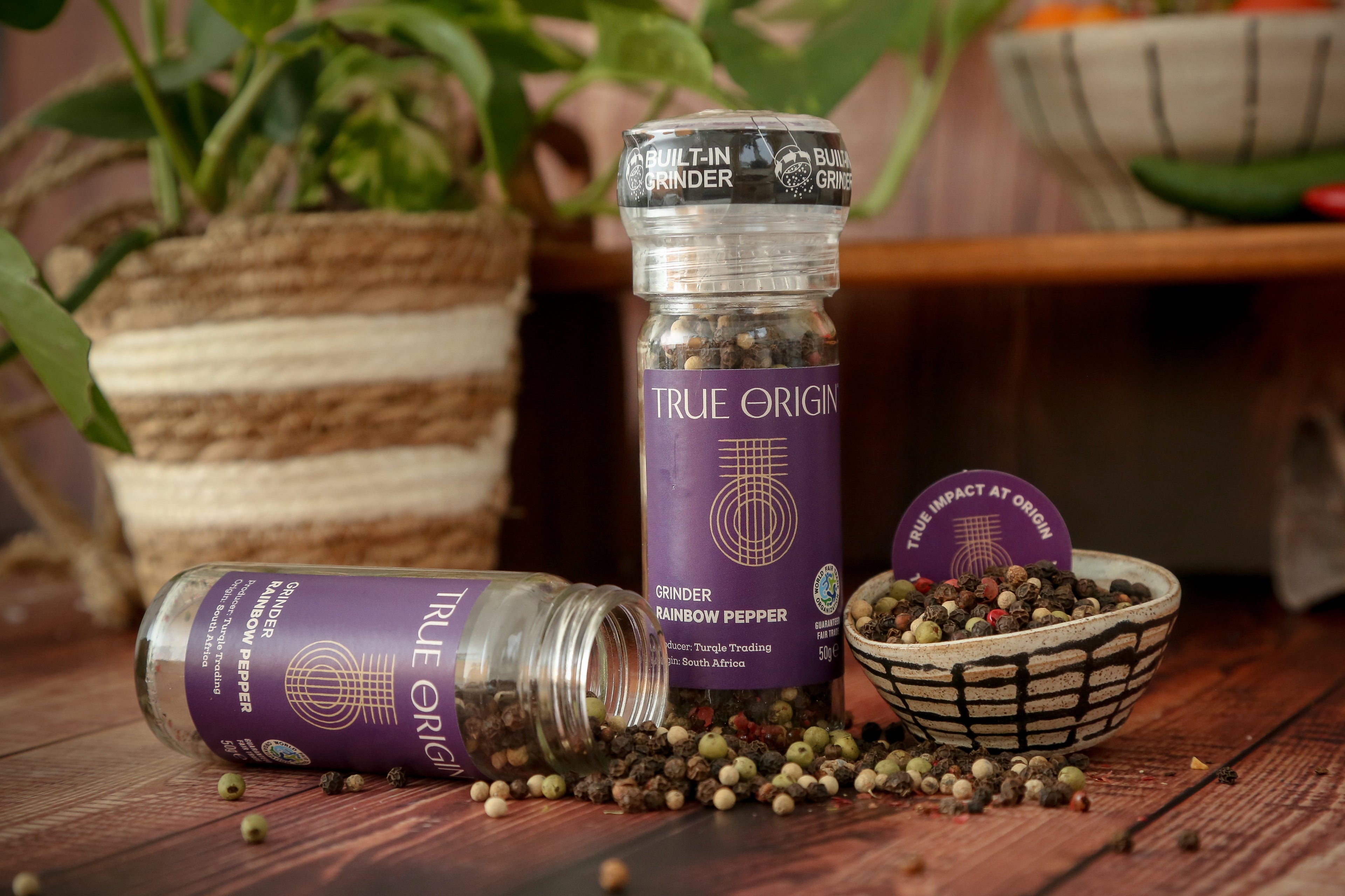 A clear glass pepper grinder with a built-in mechanism, containing a mix of black, white, green, and red pepper grains. The label shows 'True Origin' and indicates the product is a Rainbow Pepper Grinder, 50g size.