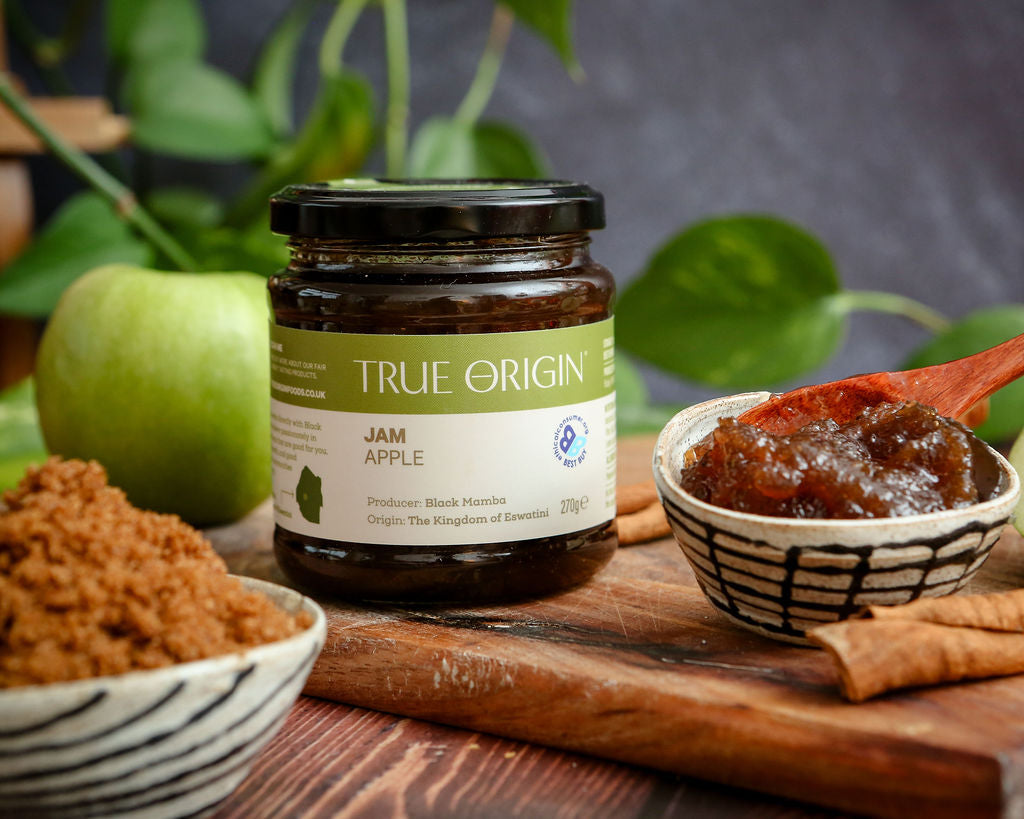 Jar of True Origin apple jam on a wooden board with apples and cinnamon sticks.