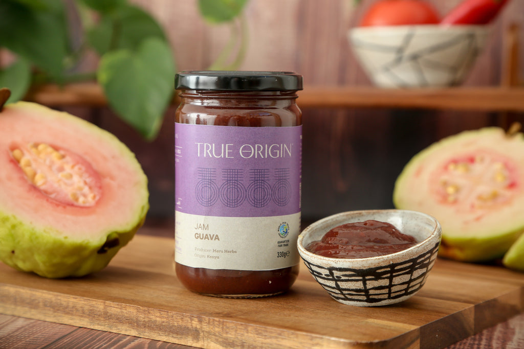 A jar of guava jam with a label that reads 'TRUE ORIGIN' and 'JAM GUAVA 330G'.  It is sitting on a wooden surface with a bowl of guava jam and some cut fresh guavas.