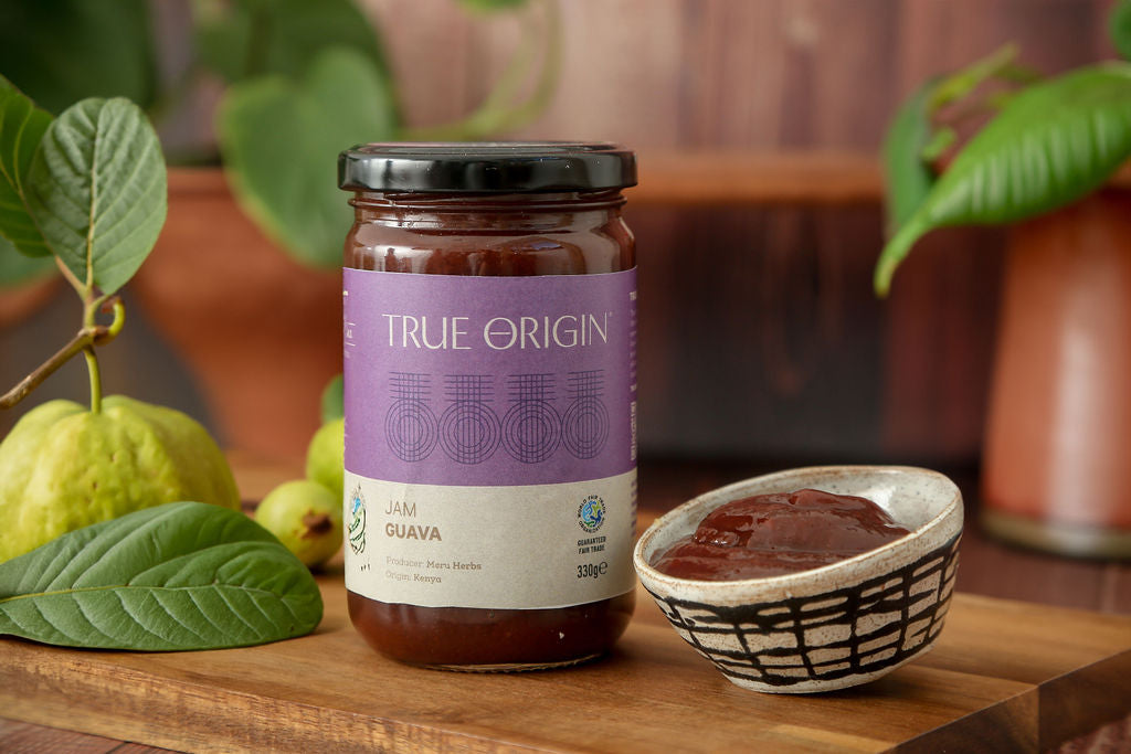 A jar of True Origin Guava Jam with a lilac and white label next to a fresh guava and a bowl of guava jam.