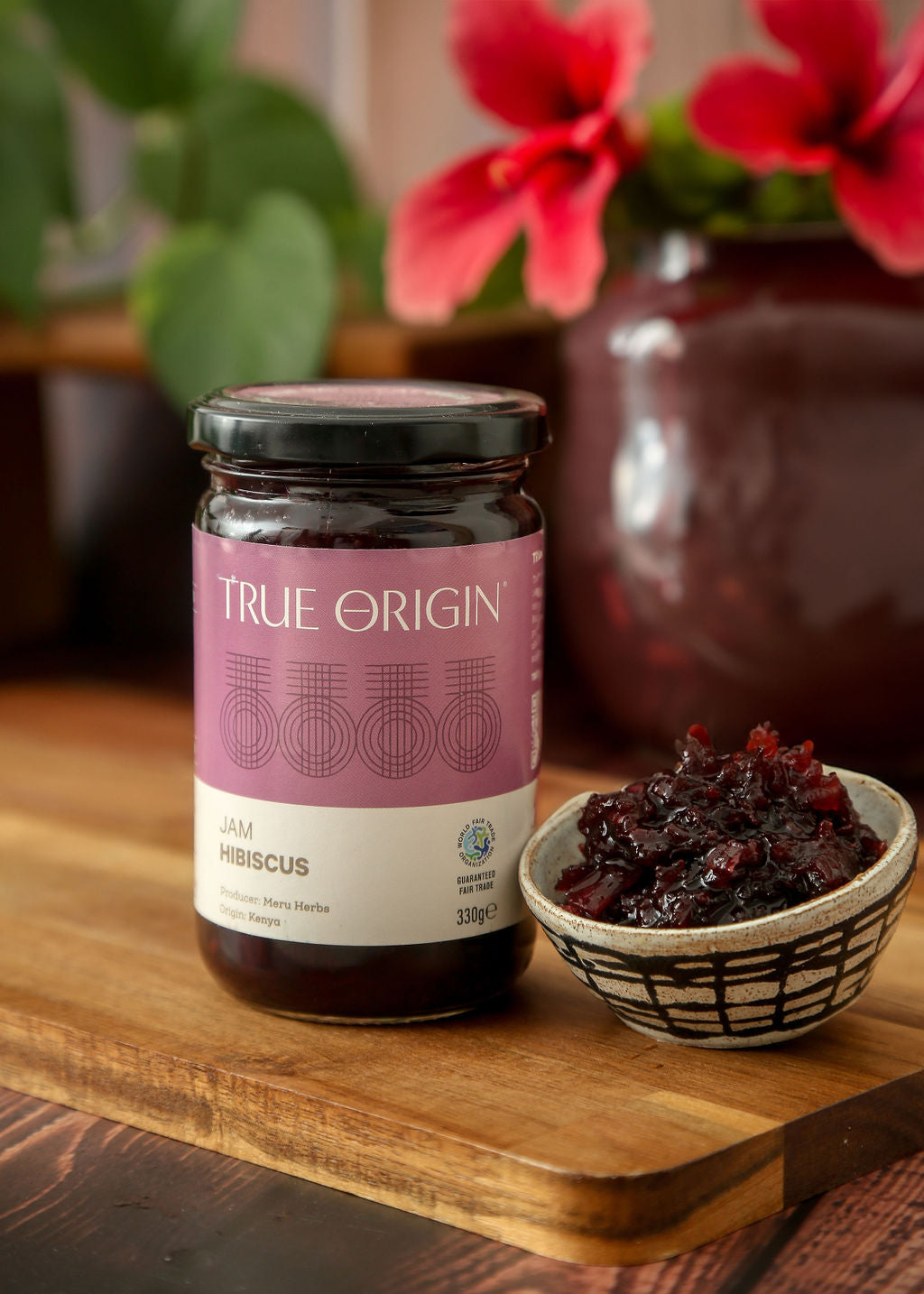 A glass jar of True Origin Hibiscus Jam on a wooden chopping board next a ceramic bowl filled with hibiscus jam from Kenya.