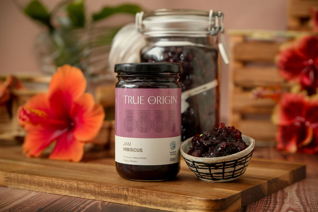 A glass jar of True Origin Hibiscus Jam on a wooden chopping board next a ceramic bowl filled with hibiscus jam from Kenya.  There are some flowers and a glass container in the background.
