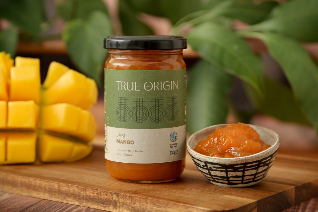 A jar of fair trade True Origin Mango Jam from Kenya.  It has a green and white label and a black lid.  It is sitting on a wooden surface next to bowl filled with Mango Jam and a cut fresh mango.
