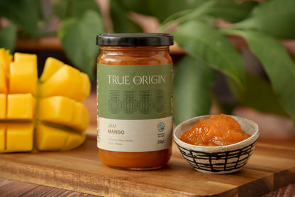 A jar of fair trade True Origin Mango Jam from Kenya.  It has a green and white label and a black lid.  It is sitting on a wooden surface next to bowl filled with Mango Jam and a cut fresh mango.