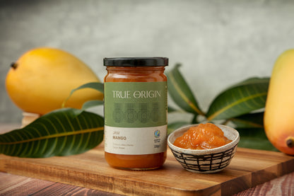 A jar of fair trade True Origin Mango Jam from Kenya.  It has a green and white label and a black lid.  It is sitting on a wooden surface next to bowl filled with Mango Jam and some fresh mangoes.