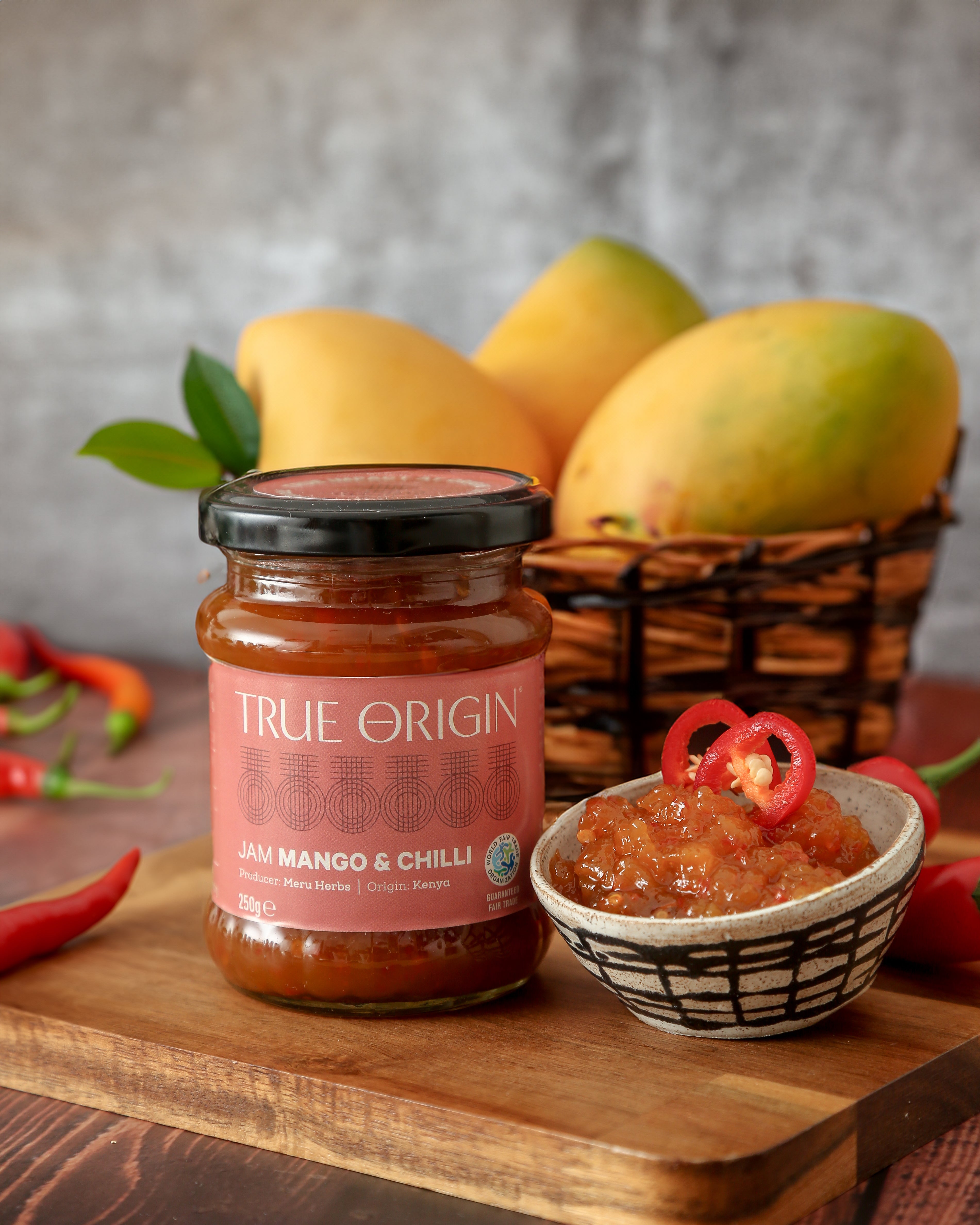 A jar of Mango & Chilli Jam with the label 'TRUE ORIGIN' displayed on it.  It is sitting next to a bowl filled with mango and chilli jam, some fresh chillies and a bowl of mangoes.