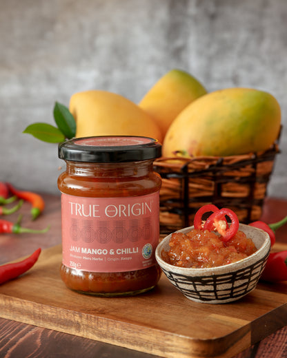 A jar of Mango & Chilli Jam with the label 'TRUE ORIGIN' displayed on it.  It is sitting next to a bowl filled with mango and chilli jam, some fresh chillies and a bowl of mangoes.