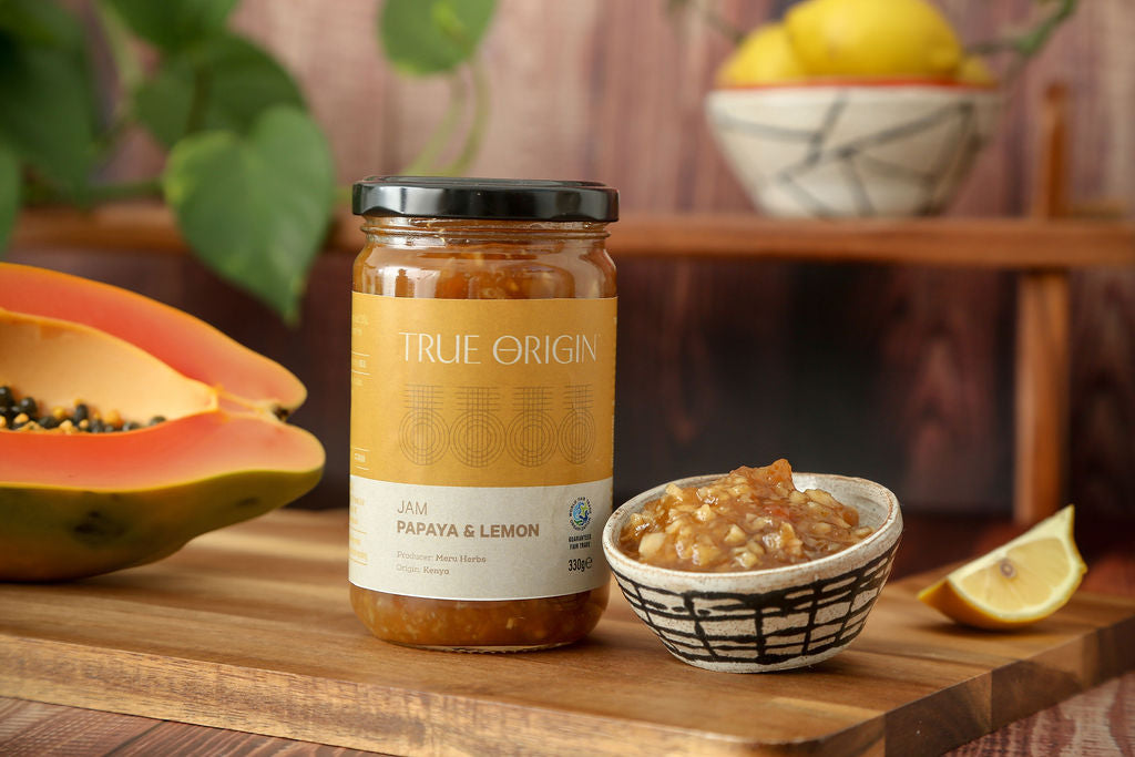A jar of True Origin papaya and lemon jam on a wooden chopping board with an open papaya and a bowl with papaya jam in it.  There is a bowl containing lemons in the background.