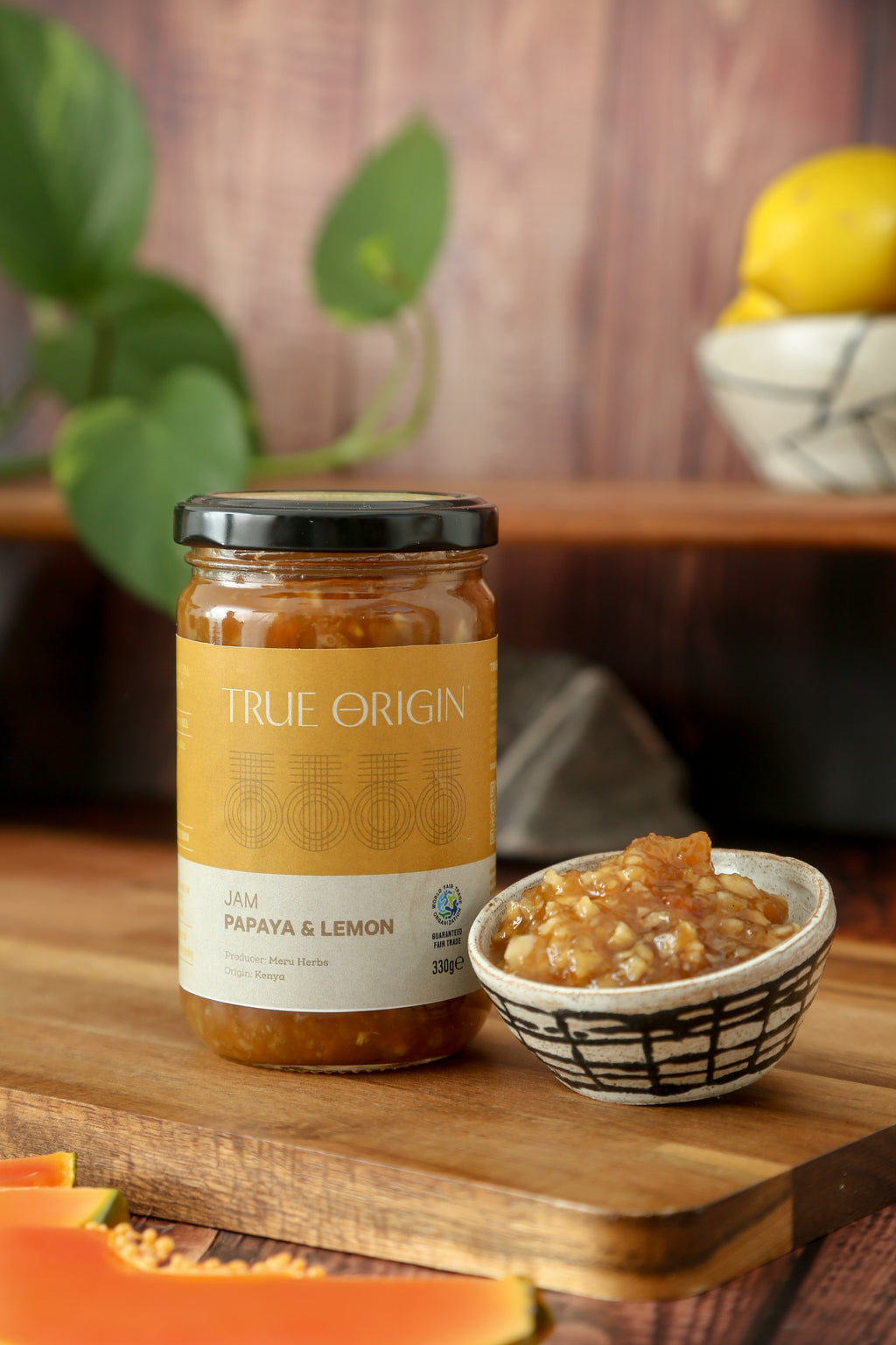 A glass jar of Papaya & Lemon Jam with a label that reads 'TRUE ORIGIN' and 'PAPAYA & LEMON JAM'. The jar is sealed with a black lid.  It is sitting on a wooden chopping board next to a bowl filled with papaya and lemon jam and some papaya slices and a bowl of lemons in the background.